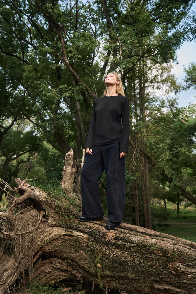 Female model standing on a fallen tree, head tilted back, eyes closed, wearing AURETEMPHE black organic cotton long sleeve t-shirt.