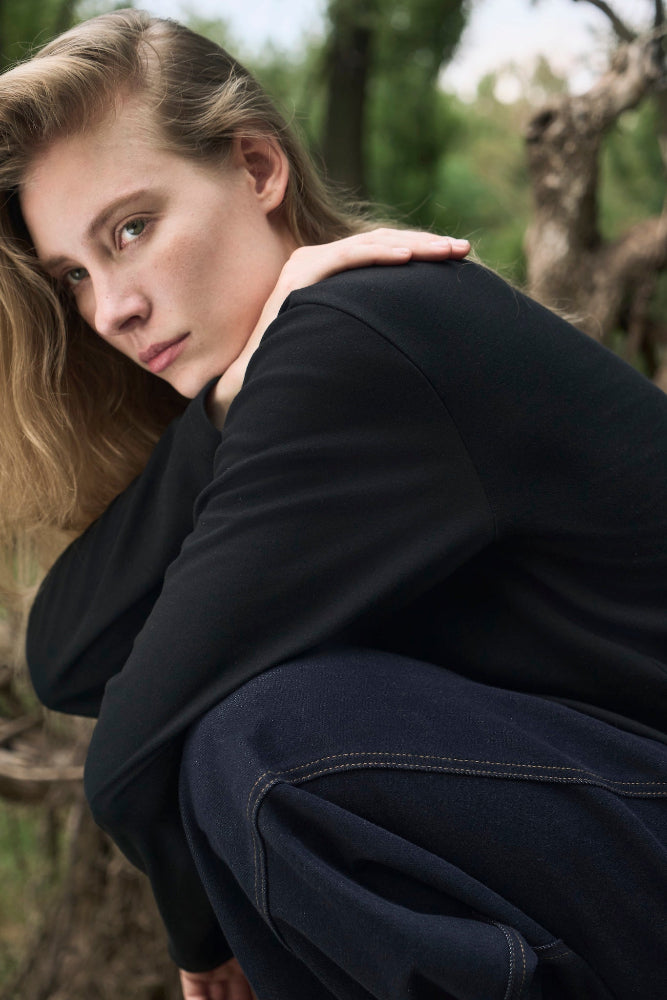 Female model crouching outdoors in nature, wearing AURETEMPHE black organic cotton long sleeve t-shirt.
