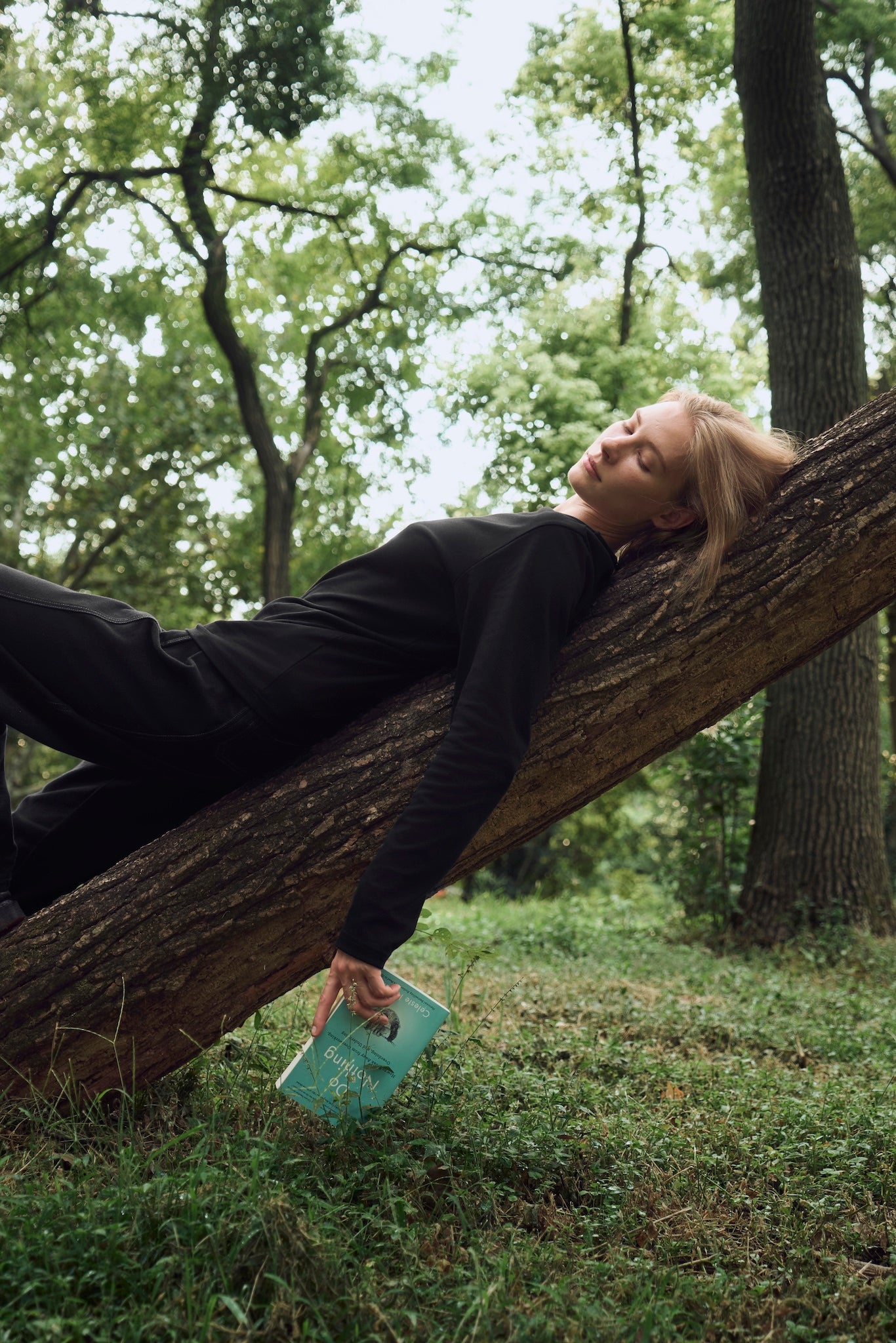 Female model resting relaxed on a fallen tree, enjoying a quiet moment in nature, wearing AURETEMPHE black organic cotton long sleeve t-shirt.