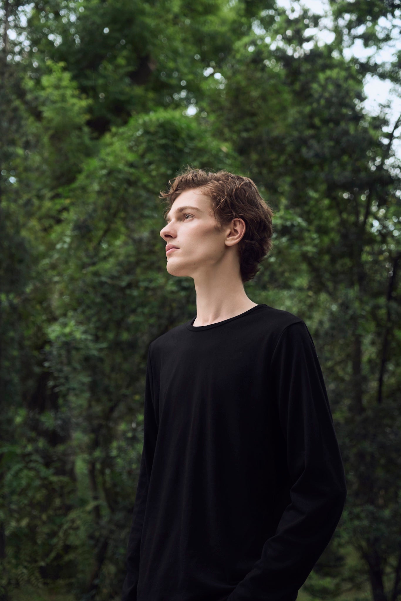 Male model exploring a forest clearing freely, with a longing gaze, wearing AURETEMPHE black organic cotton long sleeve t-shirt.