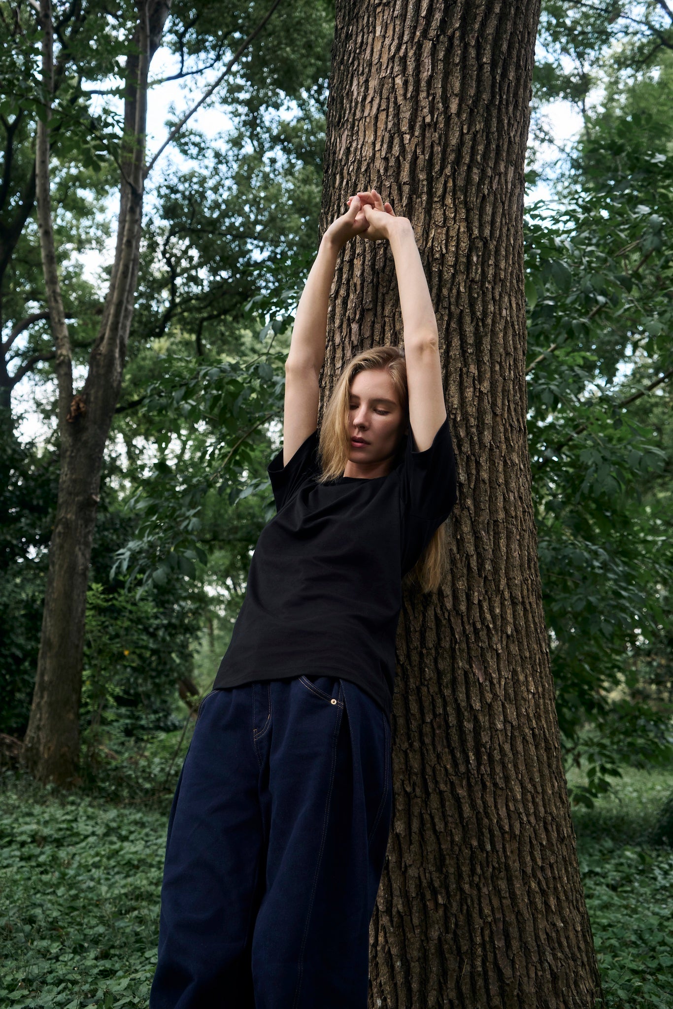Female model leaning against a tree, stretching, wearing AURETEMPHE black organic cotton short sleeve t-shirt.