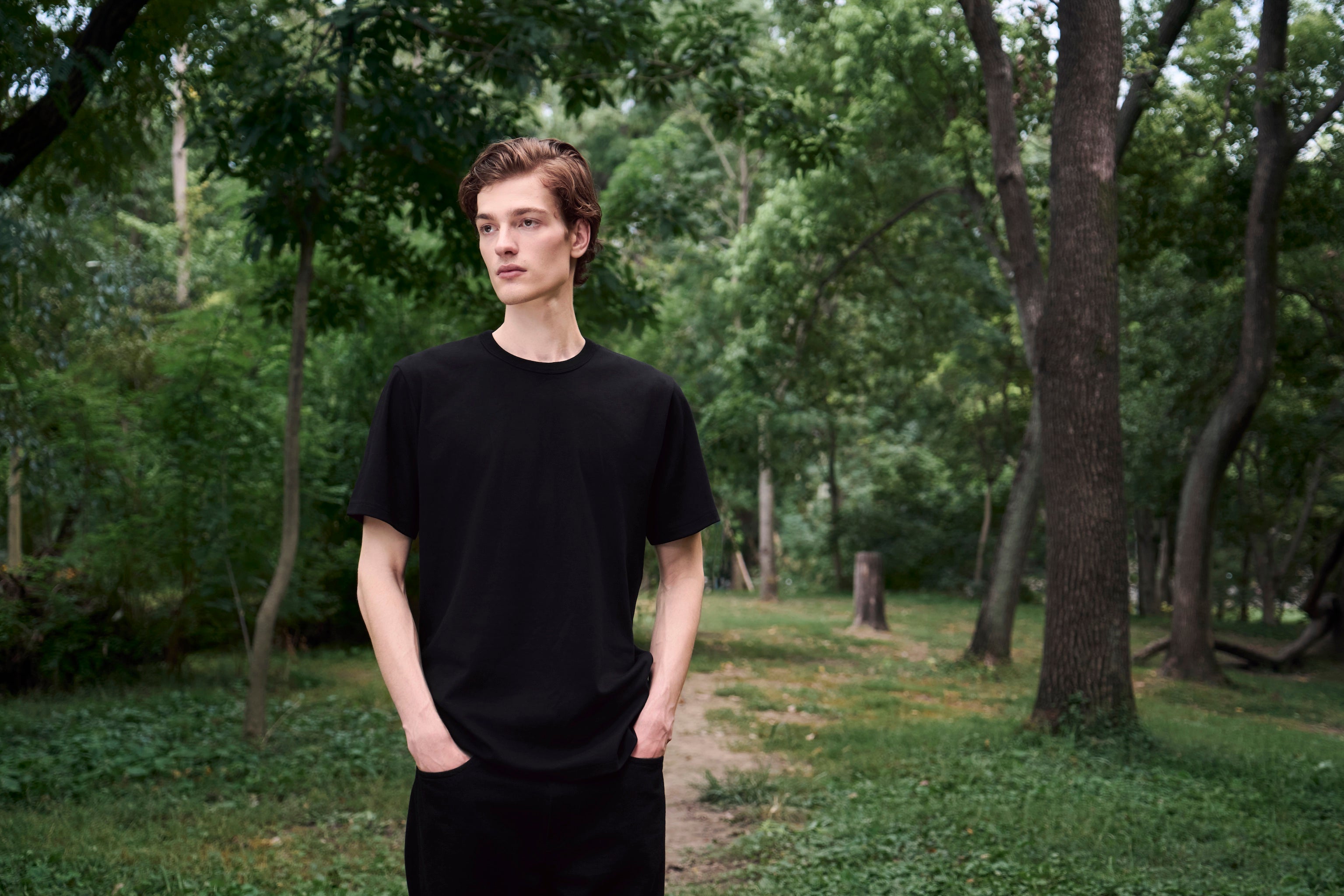 Male model standing still on a forest path with hands in pockets, wearing AURETEMPHE black organic cotton short sleeve t-shirt.