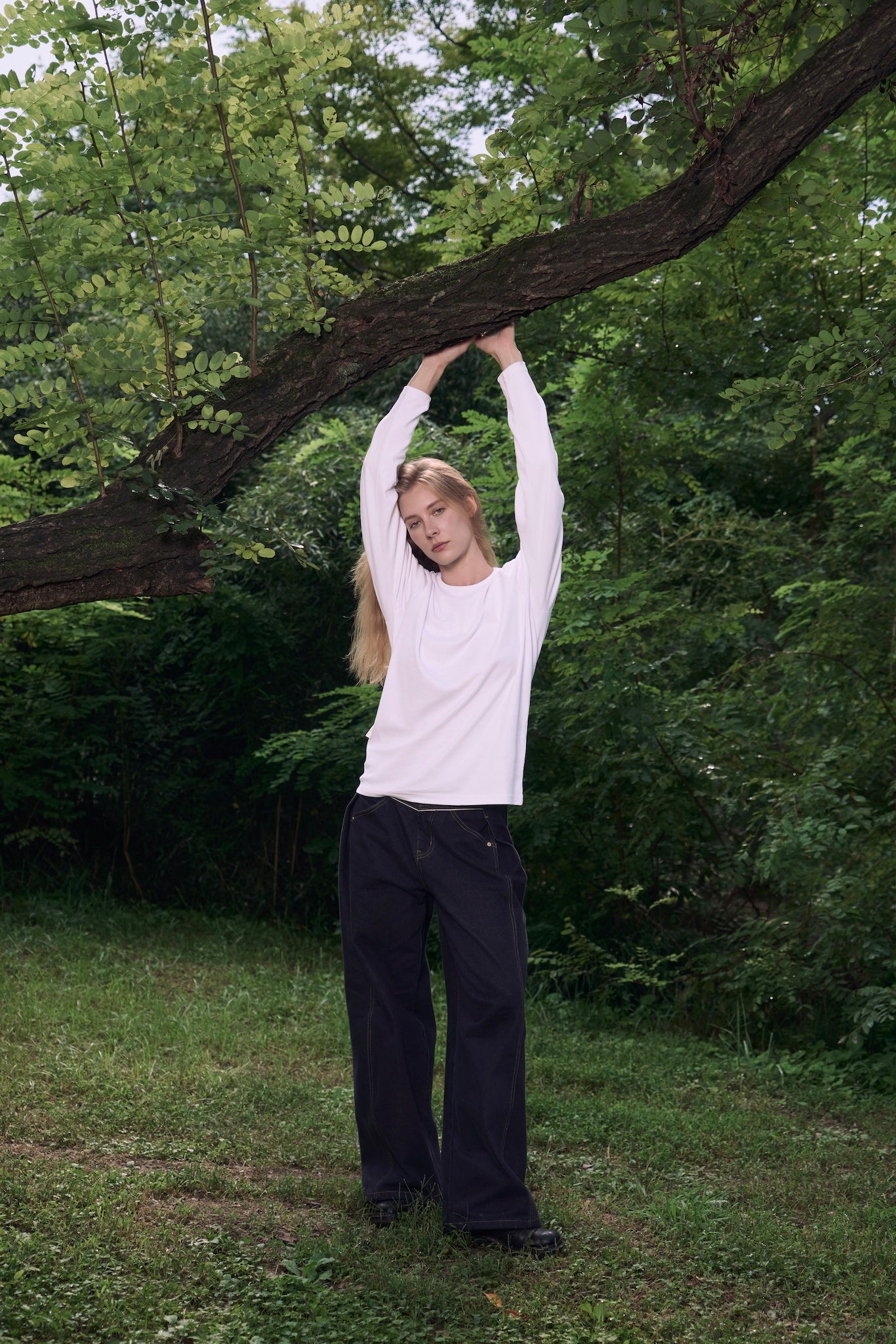 Female model with hands resting on tree branches, wearing AURETEMPHE white organic cotton long sleeve t-shirt.