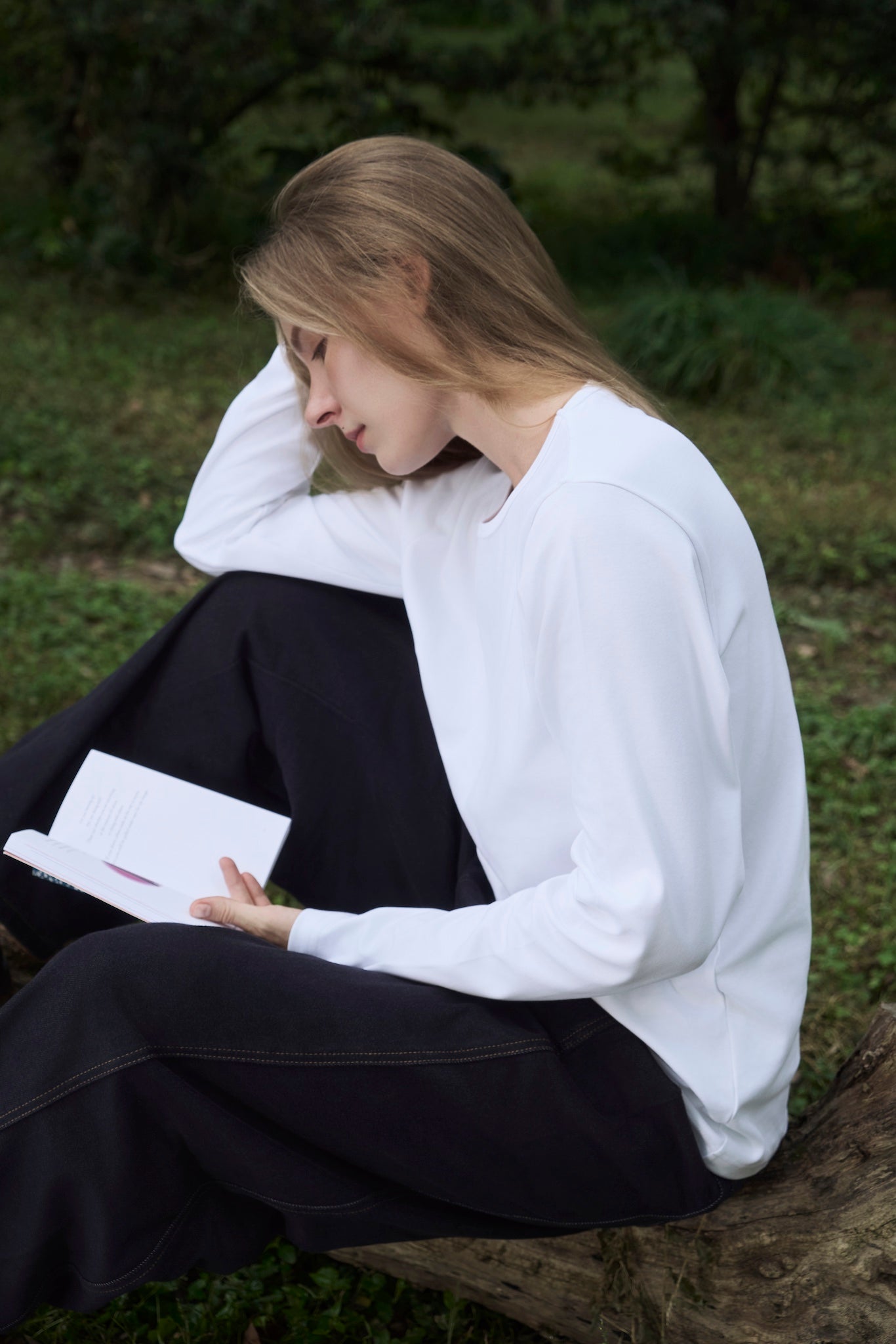 Female model reading in a forest clearing, wearing AURETEMPHE white organic cotton long sleeve t-shirt.