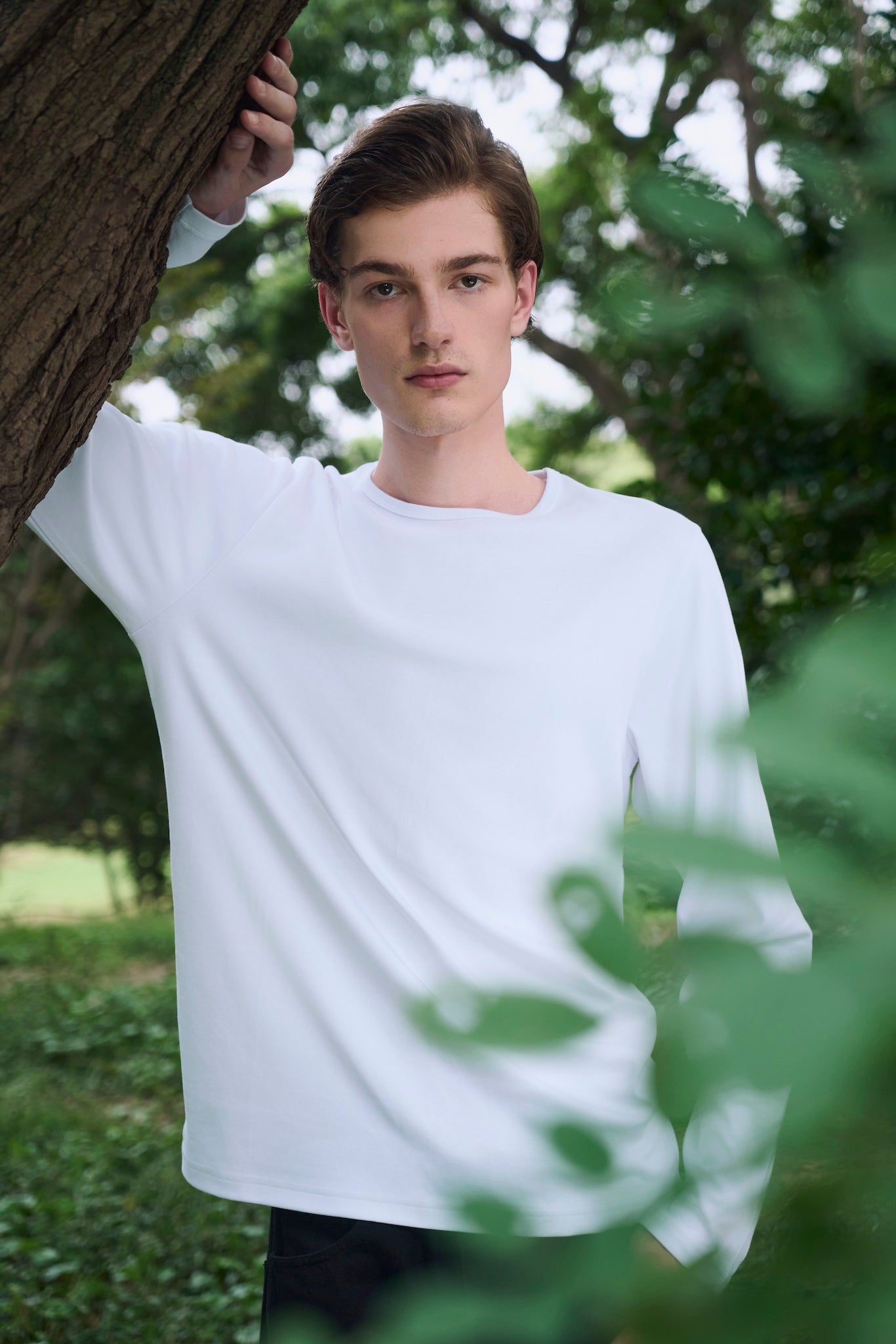 Male model resting one hand on a tree trunk, wearing AURETEMPHE white organic cotton long sleeve t-shirt.
