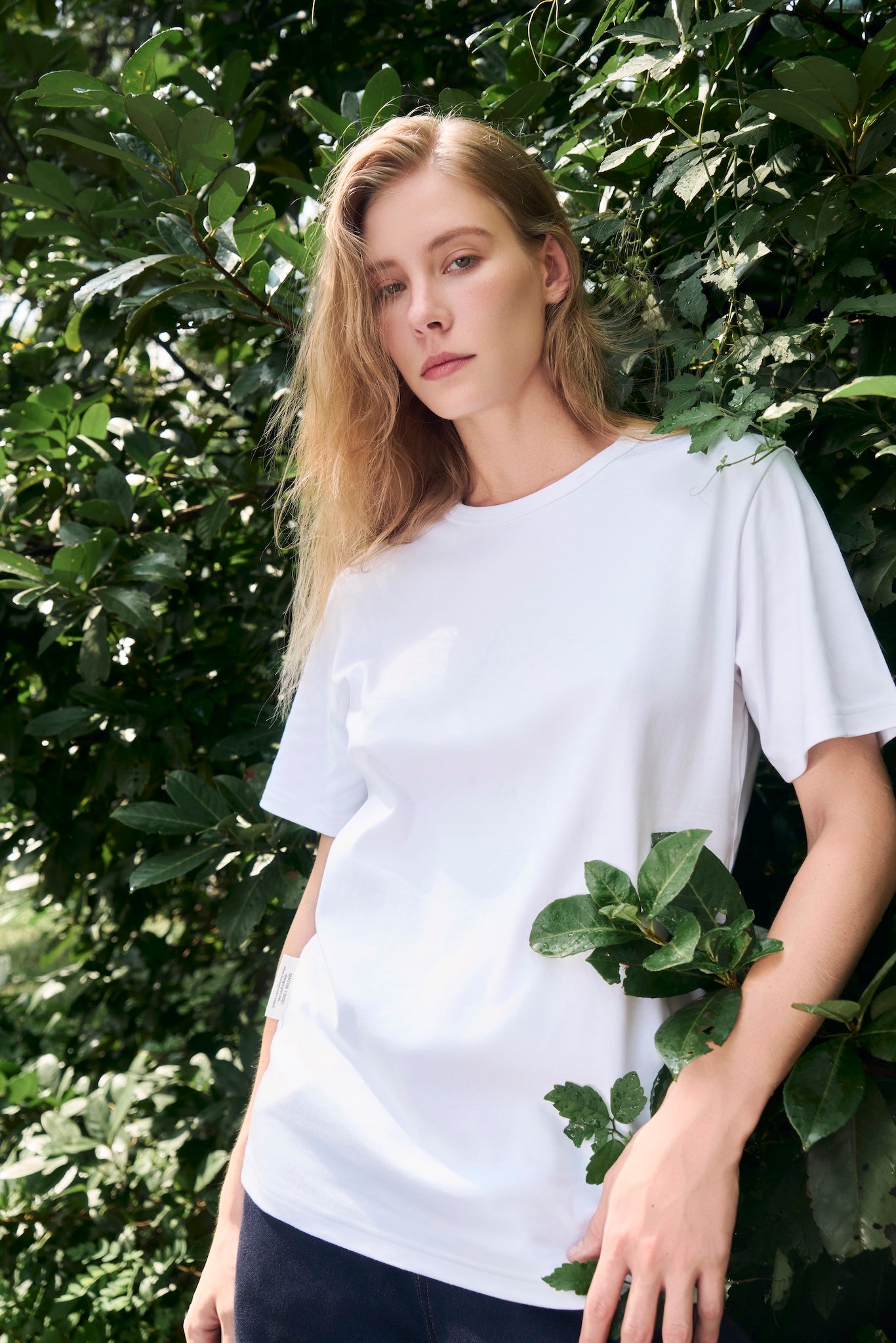 Female model standing at the edge of foliage with sunlight streaming down, wearing AURETEMPHE white organic cotton short sleeve t-shirt.