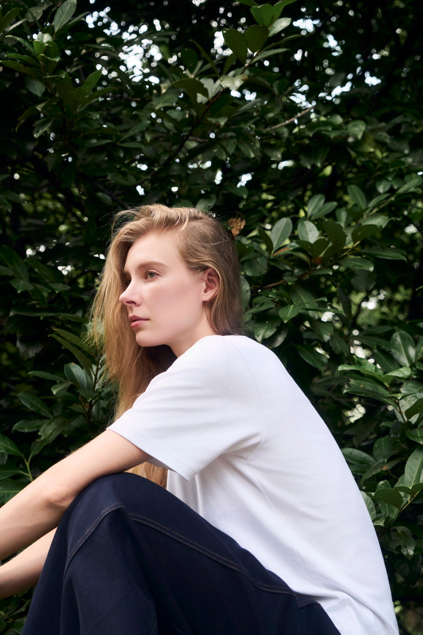 Female model crouching near foliage, wearing AURETEMPHE white organic cotton short sleeve t-shirt.